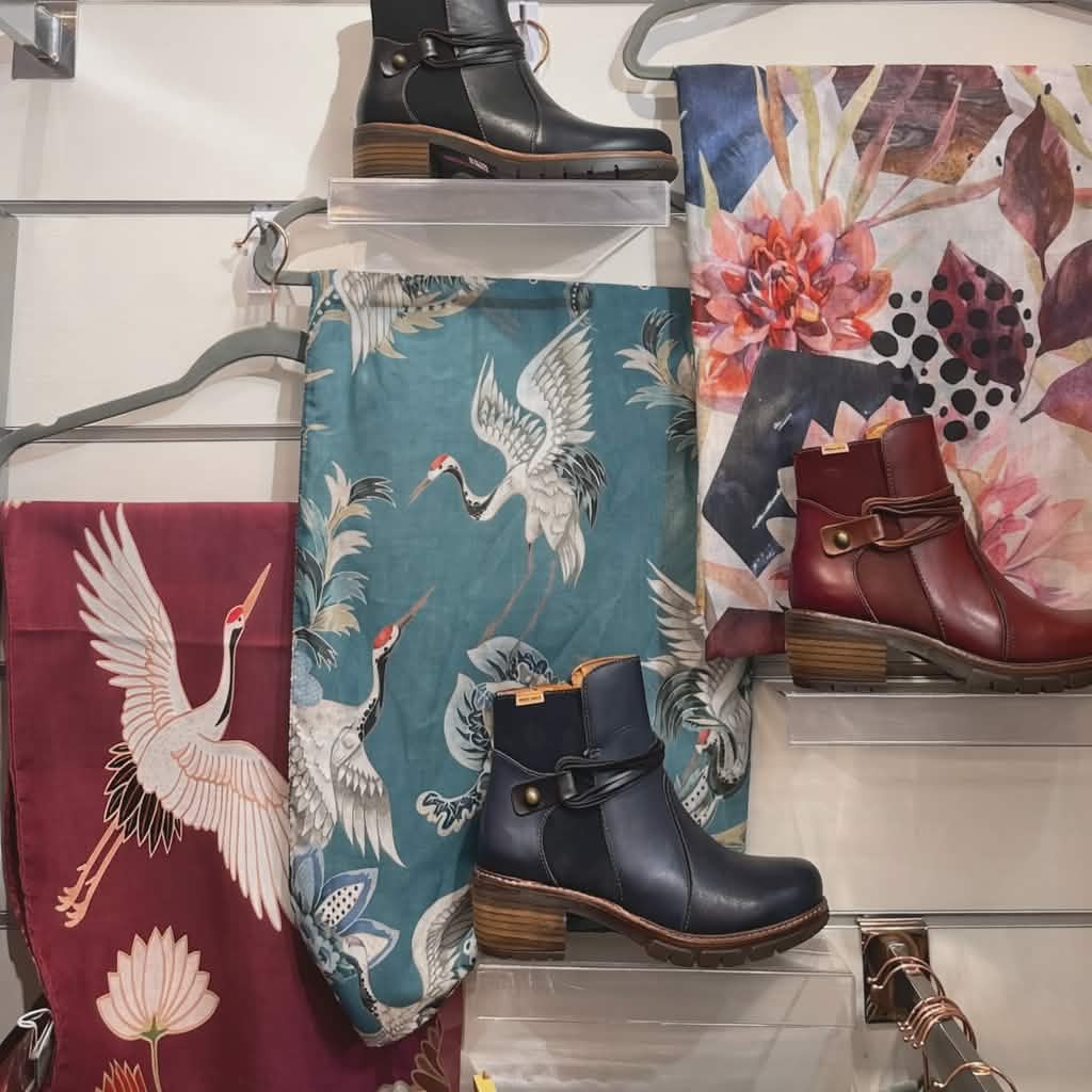 Display of ankle boots in Grants Pass, Oregon on clear shelves with three patterned scarves in teal, burgundy, and floral designs hanging on a white slatwall, creating a coordinated boutique feature.